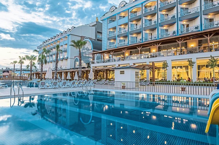 Hotelgebäude mit mehreren Stockwerken und Balkonen neben einem großen Außenpool mit Liegestühlen und Sonnenschirmen. Hotelgebäude mit mehreren Stockwerken und Balkonen neben einem großen Außenpool mit Liegestühlen und Sonnenschirmen.