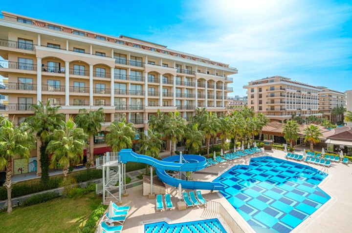 Großes Hotelgebäude mit mehreren Balkonen, davor ein Pool mit blauen Fliesen, Wasserrutsche und Liegestühlen, umgeben von Palmen. Großes Hotelgebäude mit mehreren Balkonen, davor ein Pool mit blauen Fliesen, Wasserrutsche und Liegestühlen, umgeben von Palmen.
