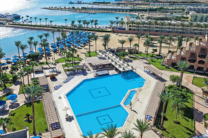 Luftaufnahme eines großen Hotelpools mit Liegen und Sonnenschirmen, umgeben von Palmen und angrenzendem Strand mit Meerblick. Luftaufnahme eines großen Hotelpools mit Liegen und Sonnenschirmen, umgeben von Palmen und angrenzendem Strand mit Meerblick.