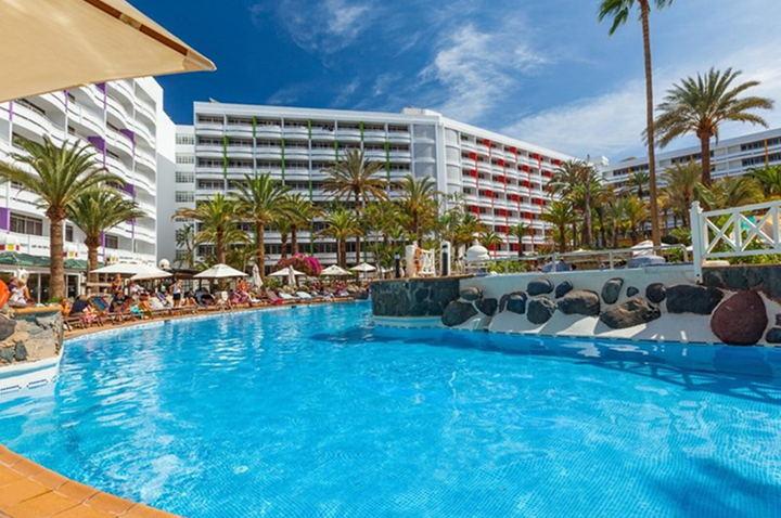 Großer Außenpool mit Sonnenschirmen und Liegestühlen vor einem mehrstöckigen Hotelgebäude und Palmen unter blauem Himmel. Großer Außenpool mit Sonnenschirmen und Liegestühlen vor einem mehrstöckigen Hotelgebäude und Palmen unter blauem Himmel.