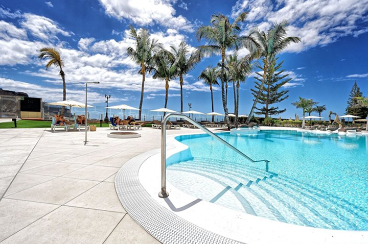 Pool mit Treppe und Edelstahlgeländer umgeben von Liegestühlen, Sonnenschirmen und Palmen unter blauem Himmel. Pool mit Treppe und Edelstahlgeländer umgeben von Liegestühlen, Sonnenschirmen und Palmen unter blauem Himmel.