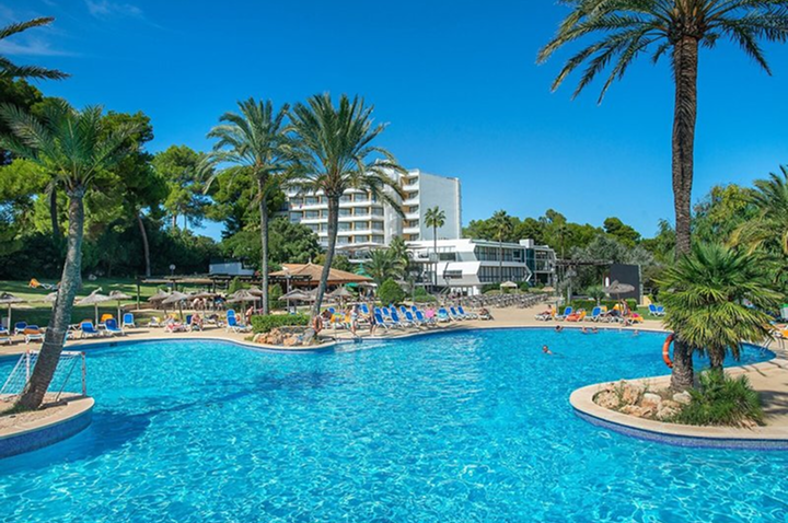 Großer Pool mit Liegestühlen und Palmen vor einem mehrstöckigen Hotelgebäude unter blauem Himmel. Großer Pool mit Liegestühlen und Palmen vor einem mehrstöckigen Hotelgebäude unter blauem Himmel.